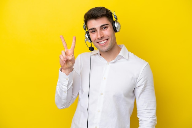 Homem caucasiano de telemarketing trabalhando com um fone de ouvido isolado em fundo amarelo sorrindo e mostrando sinal de vitória