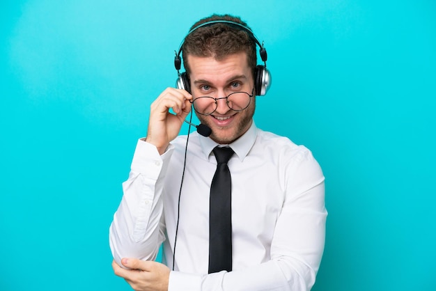Homem caucasiano de telemarketing trabalhando com um fone de ouvido isolado em fundo azul com óculos e feliz