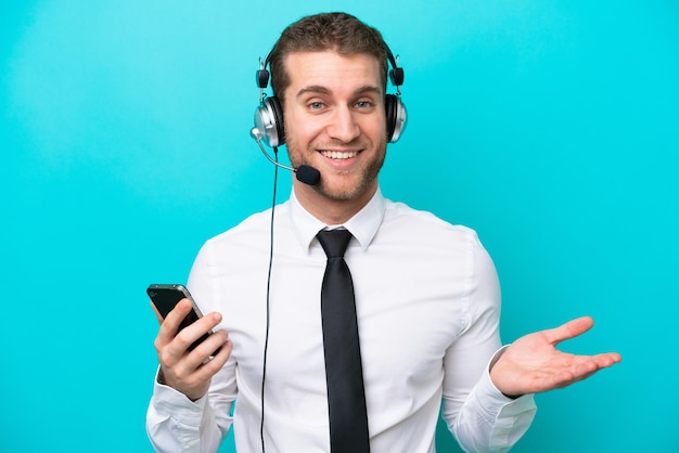 Homem caucasiano de telemarketing trabalhando com um fone de ouvido isolado em fundo azul, mantendo uma conversa com o celular com alguém