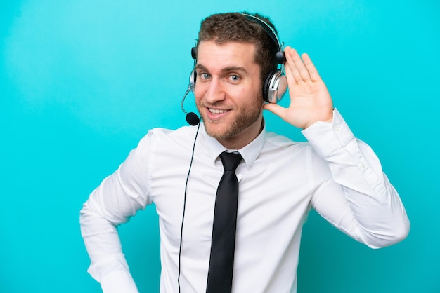 Homem caucasiano de telemarketing trabalhando com um fone de ouvido isolado em fundo azul, ouvindo algo, colocando a mão na orelha