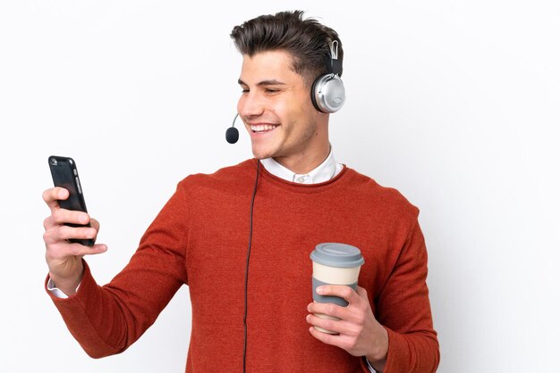 Homem caucasiano de telemarketing trabalhando com um fone de ouvido isolado no fundo branco segurando café para levar e um celular