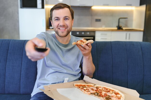 Foto homem caucasiano descansando no sofá comendo pizza e assistindo televisão conceito de estilo de vida moderno bem sucedido e relaxar depois do trabalho apartamento estúdio confortável