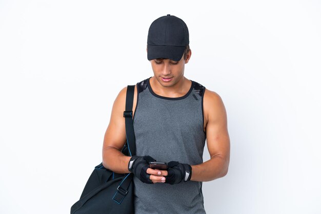 Homem caucasiano esportivo jovem com bolsa esportiva isolada em fundo branco, enviando uma mensagem com o celular