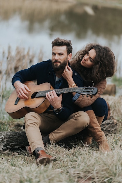 Homem caucasiano está tocando violão com uma mulher à beira do lago. Jovem casal está abraçando no dia de outono ao ar livre. Um homem barbudo e uma mulher encaracolada apaixonados.