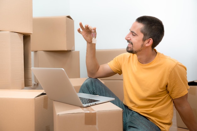 Homem caucasiano feliz com as chaves de sua nova casa, apartamento plano e laptop entre caixas.