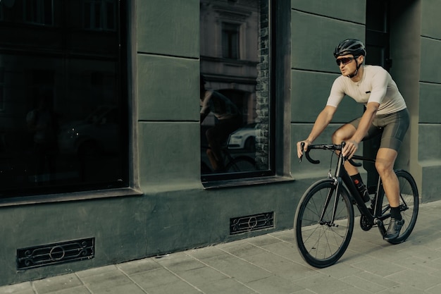 Homem caucasiano forte em capacete de segurança e óculos espelhados andando de bicicleta preta na rua da cidade Desportista ativo passando tempo livre para treino ao ar livre