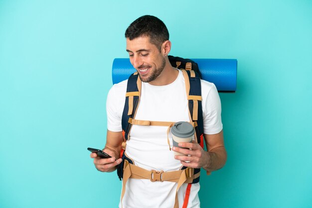 Homem caucasiano jovem alpinista com uma mochila grande isolada em fundo azul segurando café para levar e um celular