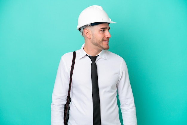 Homem caucasiano jovem arquiteto com capacete e segurando plantas isoladas em fundo azul, olhando para o lado e sorrindo