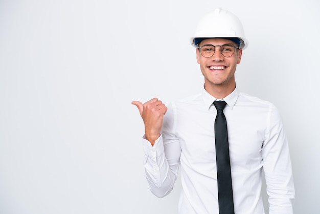 Homem caucasiano jovem arquiteto com capacete e segurando plantas isoladas no fundo branco, apontando para o lado para apresentar um produto