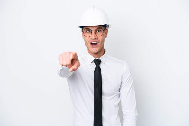 Homem caucasiano jovem arquiteto com capacete e segurando plantas isoladas no fundo branco surpreso e apontando para a frente