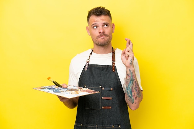 Homem caucasiano jovem artista segurando uma paleta isolada em fundo amarelo com dedos cruzando e desejando o melhor