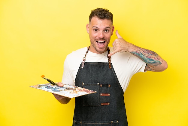 Homem caucasiano jovem artista segurando uma paleta isolada em fundo amarelo fazendo gesto de telefone Ligue-me de volta sinal