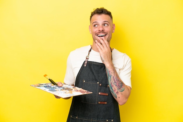 Homem caucasiano jovem artista segurando uma paleta isolada em fundo amarelo olhando para cima enquanto sorri