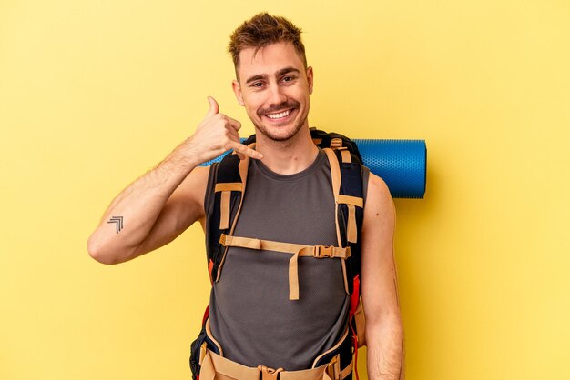 Homem caucasiano jovem caminhante isolado em fundo amarelo, mostrando um gesto de chamada de telefone móvel com os dedos.