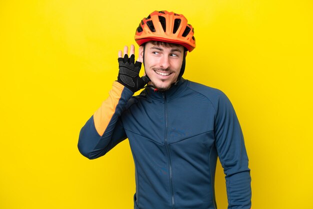 Homem caucasiano jovem ciclista isolado em fundo amarelo ouvindo algo colocando a mão na orelha