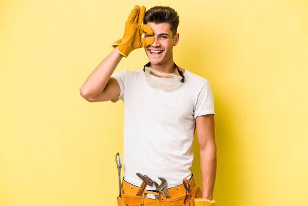 Homem caucasiano jovem eletricista isolado em fundo amarelo animado mantendo o gesto ok no olho