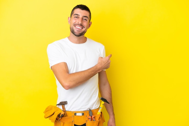 Homem caucasiano jovem eletricista isolado em fundo amarelo, apontando para trás