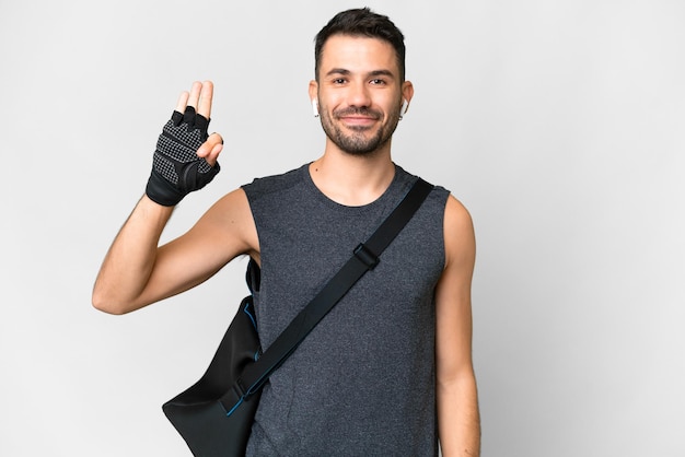 Homem caucasiano jovem esporte com bolsa esportiva sobre fundo branco isolado feliz e contando três com os dedos