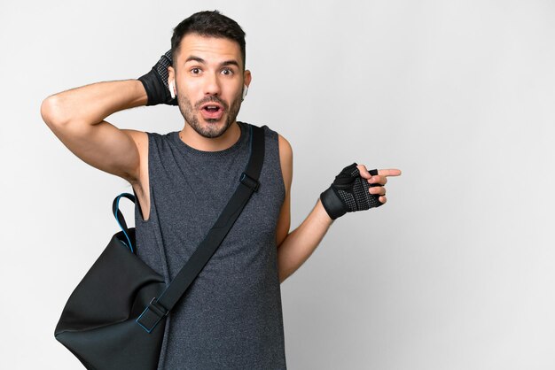 Homem caucasiano jovem esporte com bolsa esportiva sobre fundo branco isolado surpreso e apontando o dedo para o lado