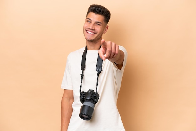 Homem caucasiano jovem fotógrafo isolado em fundo bege aponta o dedo para você com uma expressão confiante
