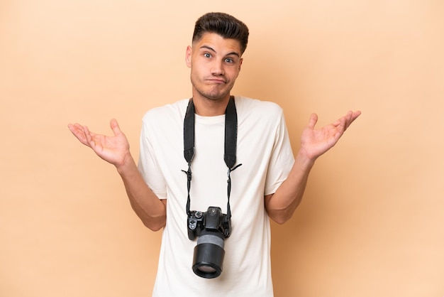 Homem caucasiano jovem fotógrafo isolado em fundo bege com dúvidas ao levantar as mãos