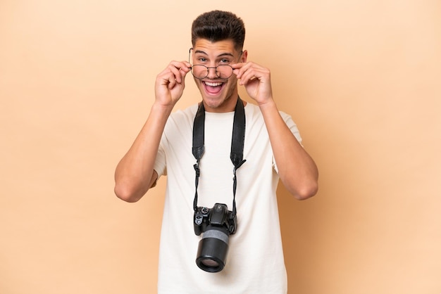 Homem caucasiano jovem fotógrafo isolado em fundo bege com óculos e surpreso