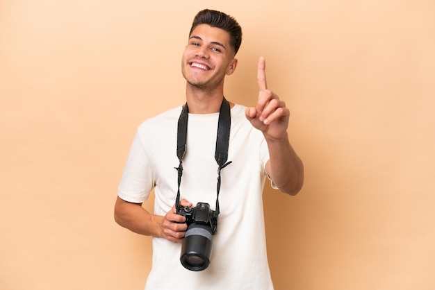 Homem caucasiano jovem fotógrafo isolado em fundo bege mostrando e levantando um dedo
