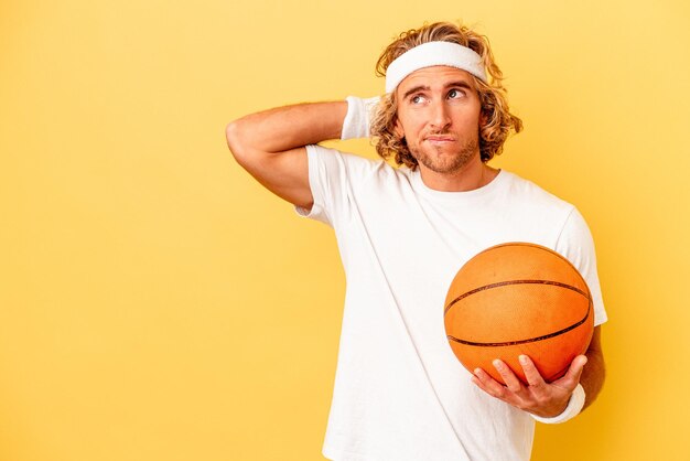 Homem caucasiano jovem jogador de basquete isolado em fundo amarelo, tocando a parte de trás da cabeça, pensando e fazendo uma escolha.