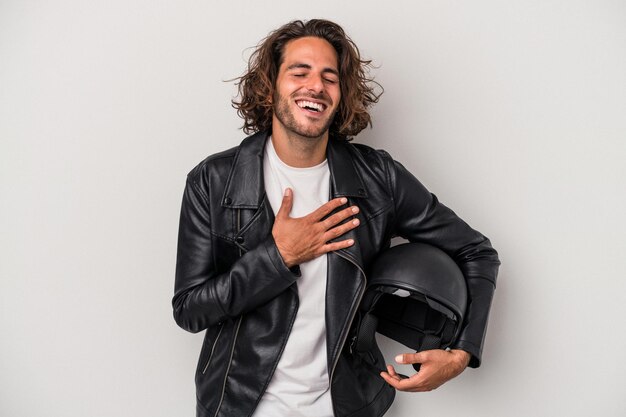 Homem caucasiano jovem motociclista segurando um capacete de moto isolado no fundo cinza ri alto, mantendo a mão no peito.