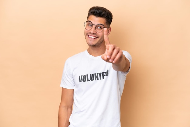 Homem caucasiano jovem voluntário isolado em fundo bege mostrando e levantando um dedo