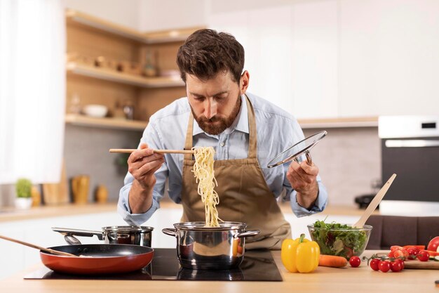 Homem caucasiano maduro bonito sério com barba no avental preparar macarrão no interior da cozinha minimalista