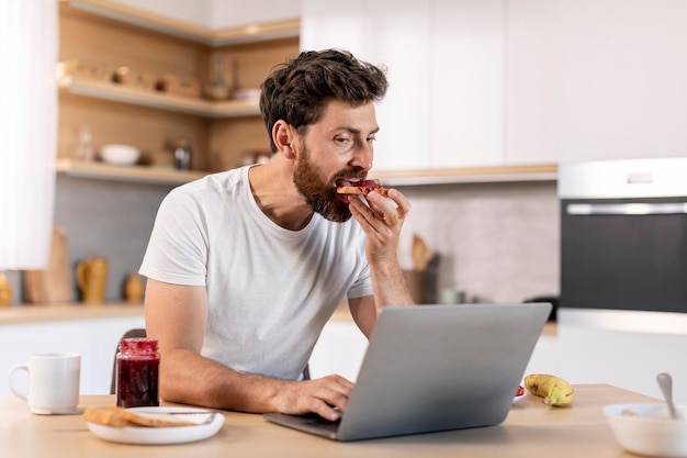 Homem caucasiano milenar ocupado com barba come sanduíche com geléia e trabalha em computador navegando na internet