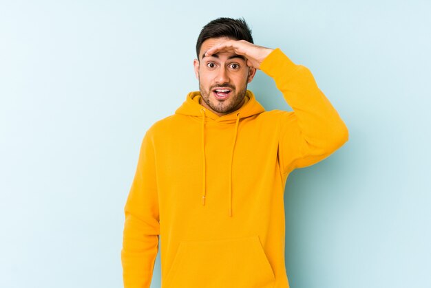 Homem caucasiano novo isolado no azul que olha longe mantendo a mão na testa.