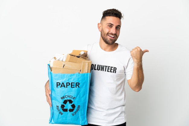 Homem caucasiano segurando um saco de reciclagem cheio de papel para reciclar isolado em uma parede branca apontando para o lado para apresentar um produto