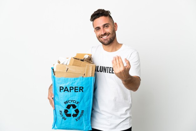 Homem caucasiano segurando uma sacola cheia de papel para reciclar isolado no fundo branco, convidando para vir com a mão. Feliz que você veio