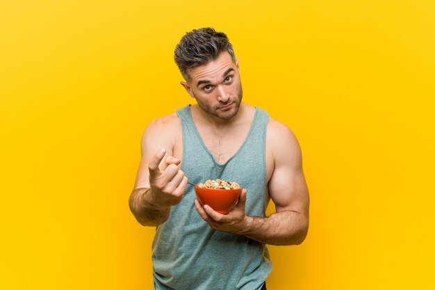 Foto homem caucasiano, segurando uma tigela de cereais, apontando com o dedo para você, como se estivesse convidando para se aproximar.