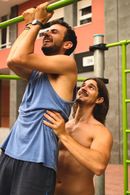 Homem caucasiano sem camisa, ajudando seu amigo a fazer uma flexão em um parque de ginástica ao ar livre