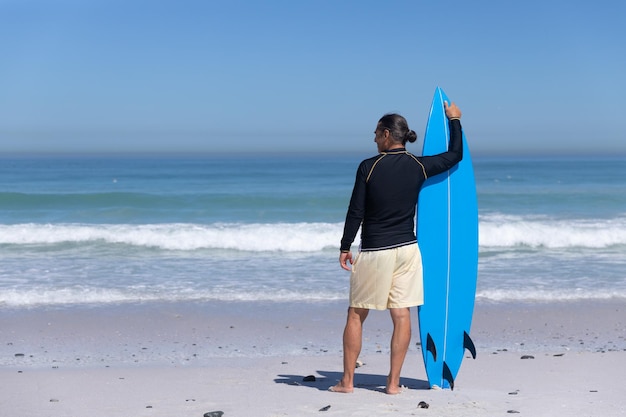 Homem caucasiano sênior aproveitando o tempo na praia em um dia ensolarado, de pé e segurando uma prancha de surf com o mar ao fundo