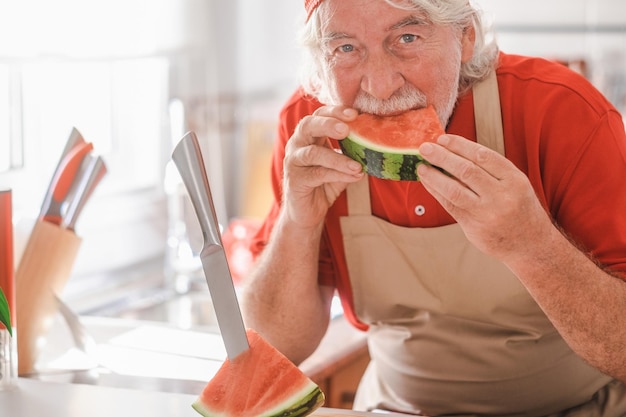 Homem caucasiano sênior na cozinha de casa enquanto come uma fatia de melancia vermelha fresca ativa sorrindo aposentado barbudo vestido com o conceito de frutas vermelhas de verão