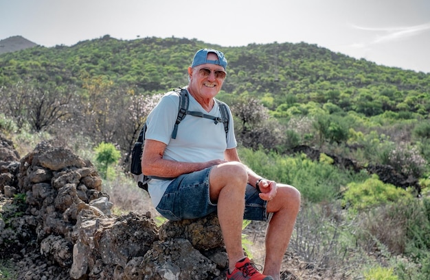 Homem caucasiano sênior sorridente com boné e mochila em excursão de montanha ao ar livre senta-se para descansar