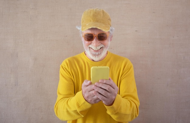 Homem caucasiano sênior sorridente em amarelo com óculos escuros e chapéu segurando um telefone inteligente olhando vídeos engraçados