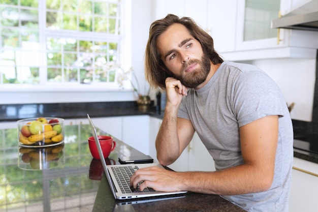 Homem caucasiano sentado na cozinha usando laptop e pensando