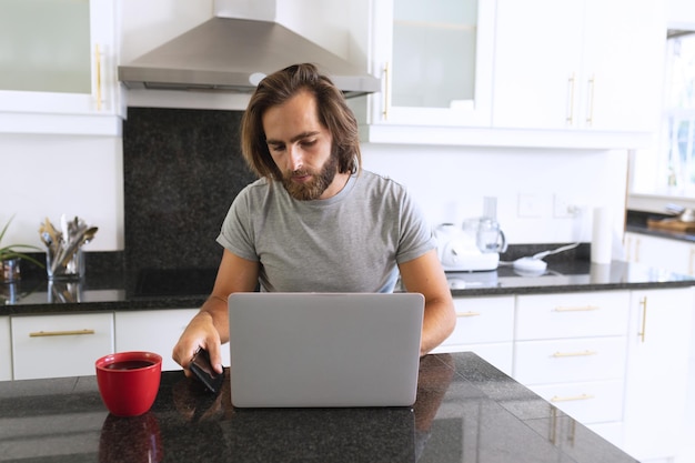 Homem caucasiano sentado na cozinha usando laptop e smartphone