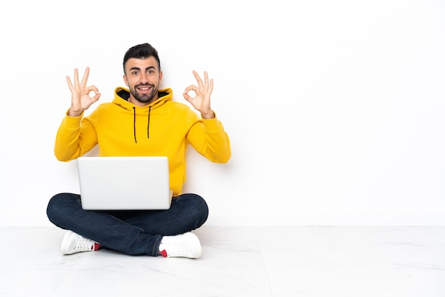 Homem caucasiano sentado no chão com seu laptop, mostrando um sinal de ok com os dedos