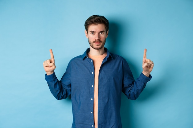 Homem caucasiano sério com barba apontando os dedos para o espaço vazio, mostrando o logotipo e olhando para a câmera, de pé sobre fundo azul.