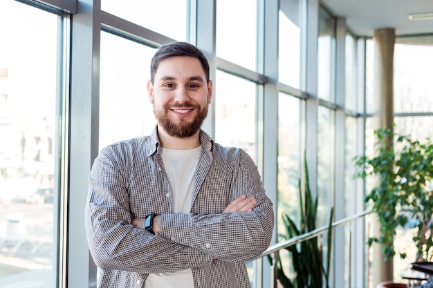 Homem caucasiano sorridente em pé perto da janela de corpo inteiro no escritório sozinho. bem-sucedido feliz barbudo jovem cruzou os braços. homem inteligente bonito empresário com camisa casual. freelancer no espaço de coworking.