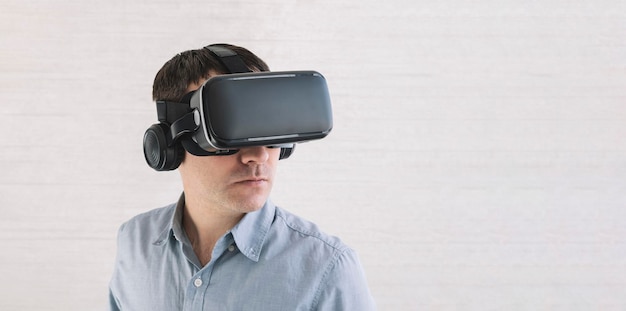 Homem caucasiano surpreso em camiseta azul usando fone de ouvido oculus rift experimentando realidade virtual enquanto joga videogame parecendo alegre em pé contra a parede branca visualização de vídeo em 360 graus