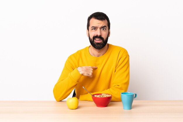 Homem caucasiano tomando café da manhã em uma mesa apontando para si mesmo.