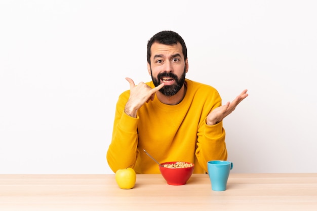 Homem caucasiano, tomando café da manhã em uma mesa, fazendo gesto de telefone e duvidando