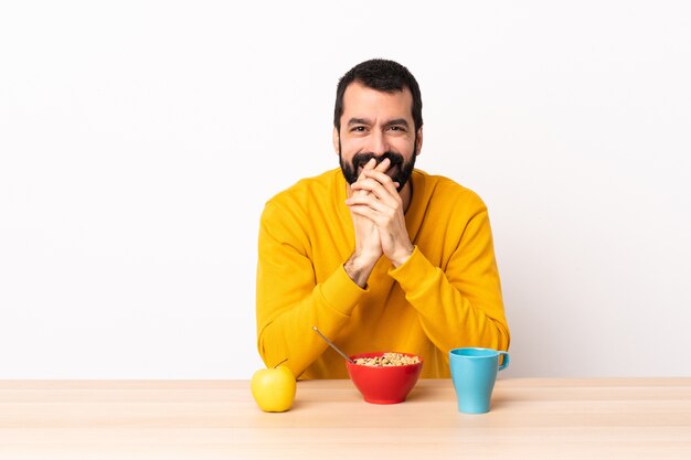 Homem caucasiano tomando café da manhã em uma mesa feliz e sorridente, cobrindo a boca com as mãos